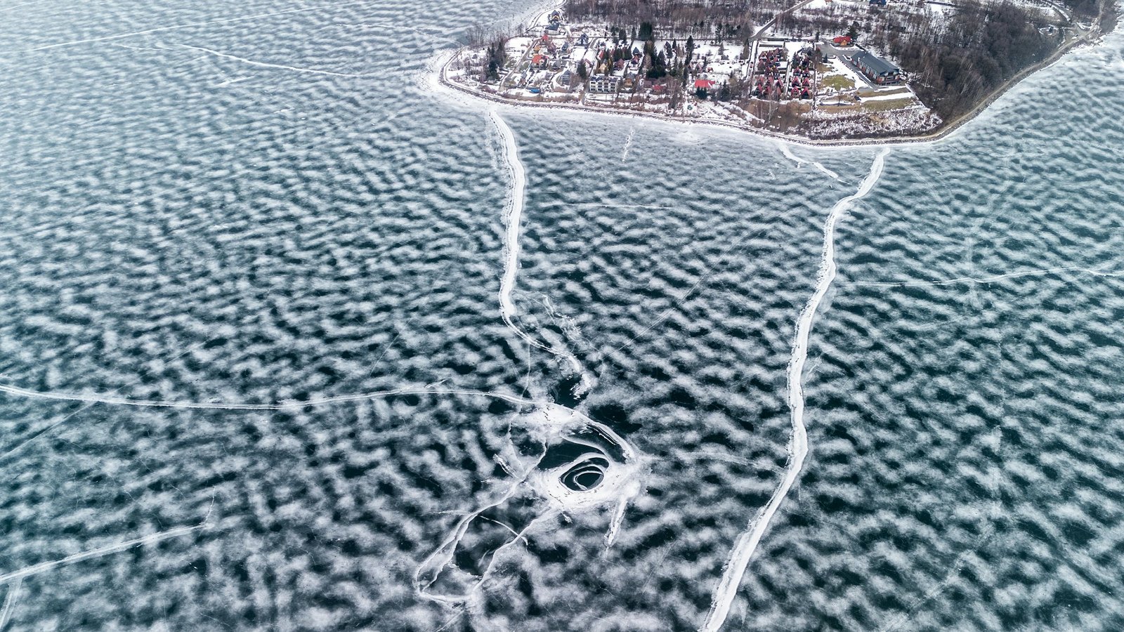 Aerial of a frozen lake with snowmobile tracks across the ice and a small lakeside community along the shore.